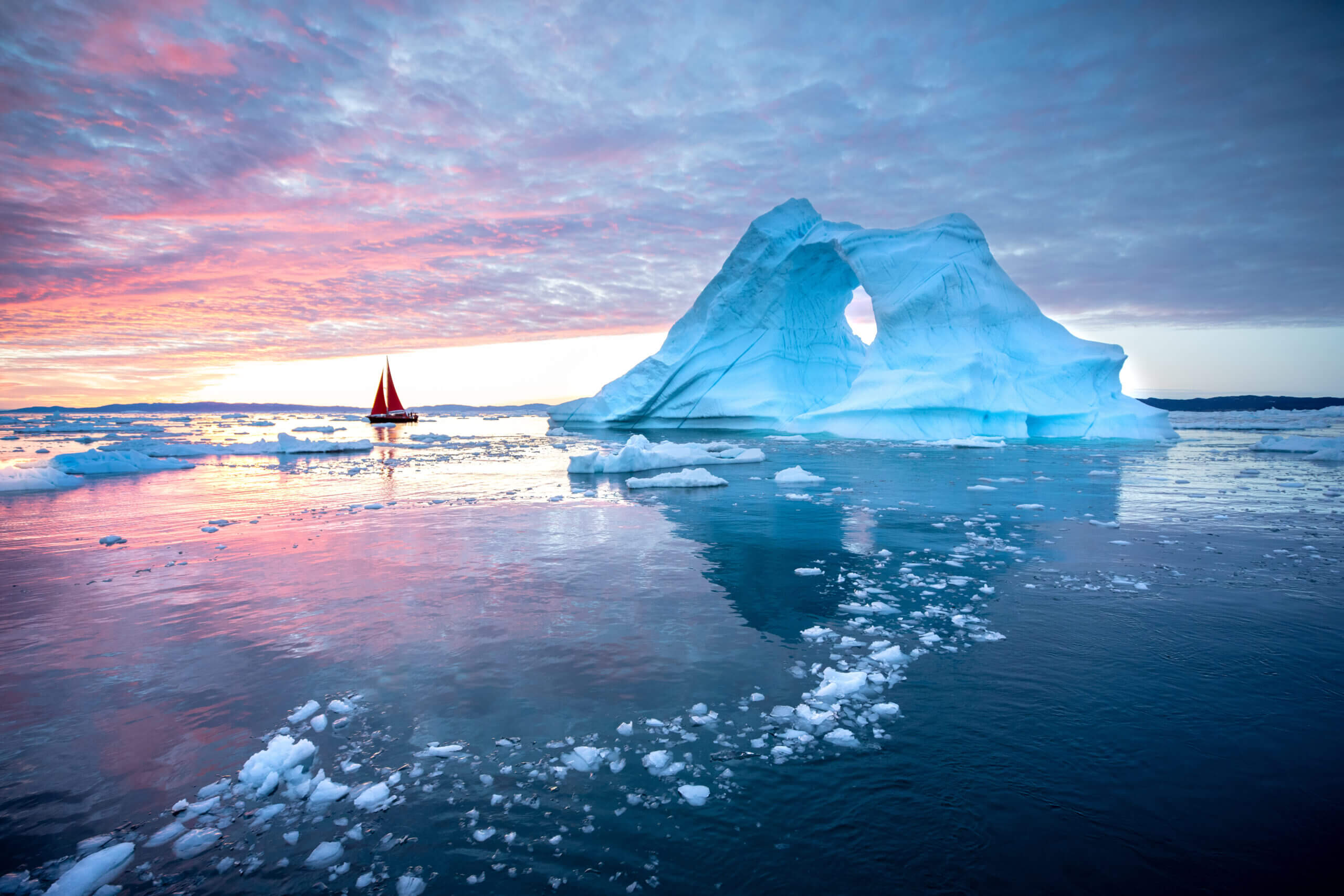 Red sailboat cruising among floating icebergs in Disko Bay during midnight sun season of polar summer