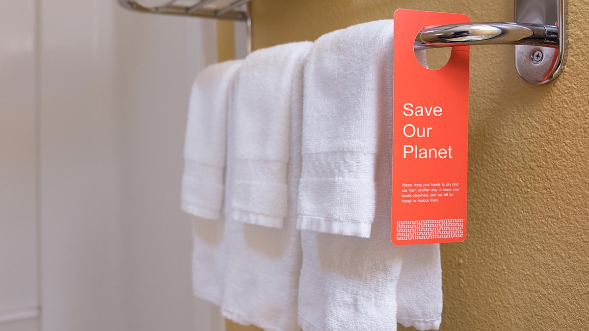 A save water sign sits on a towel rack in a hotel bathroom. Hotels use signs like this to trick travelers into doing the sustainability work for them, which is a form of greenwashing.