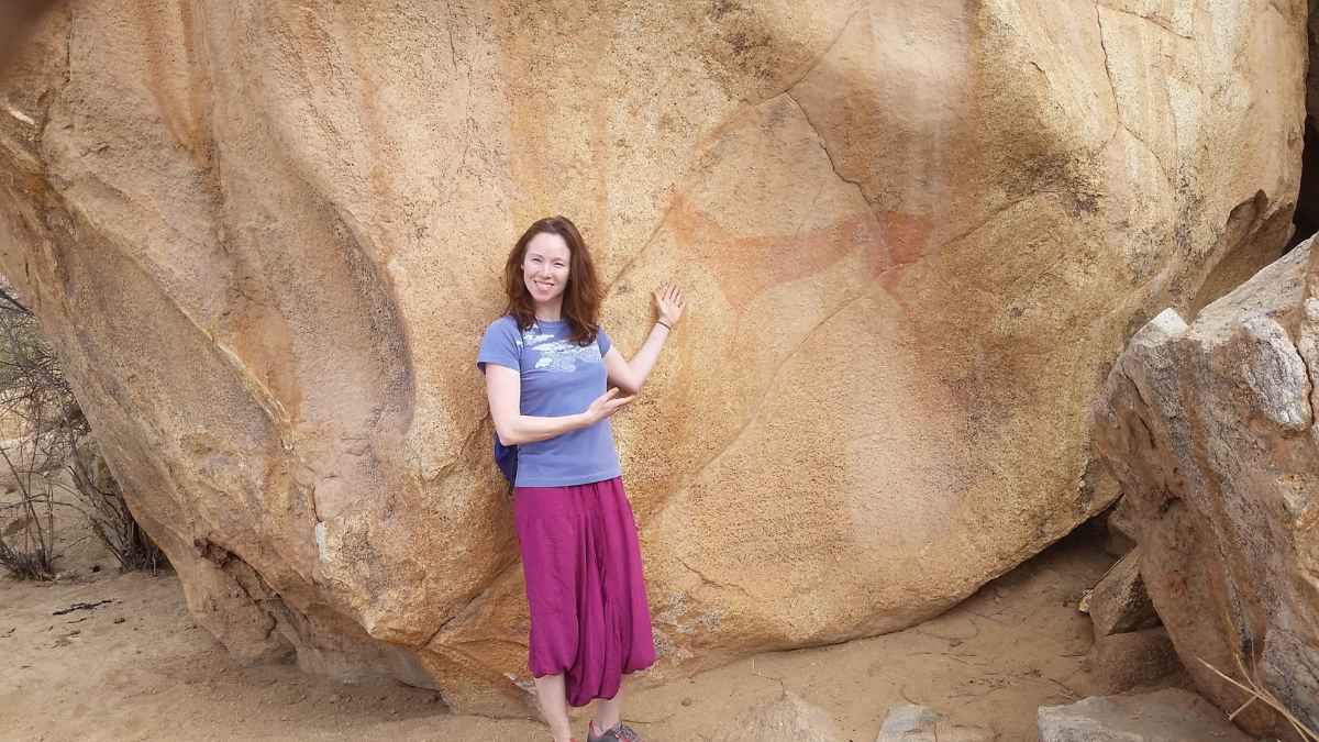 Amanda stands by and points at an ancient orange cave painting on brown rock in Namibia