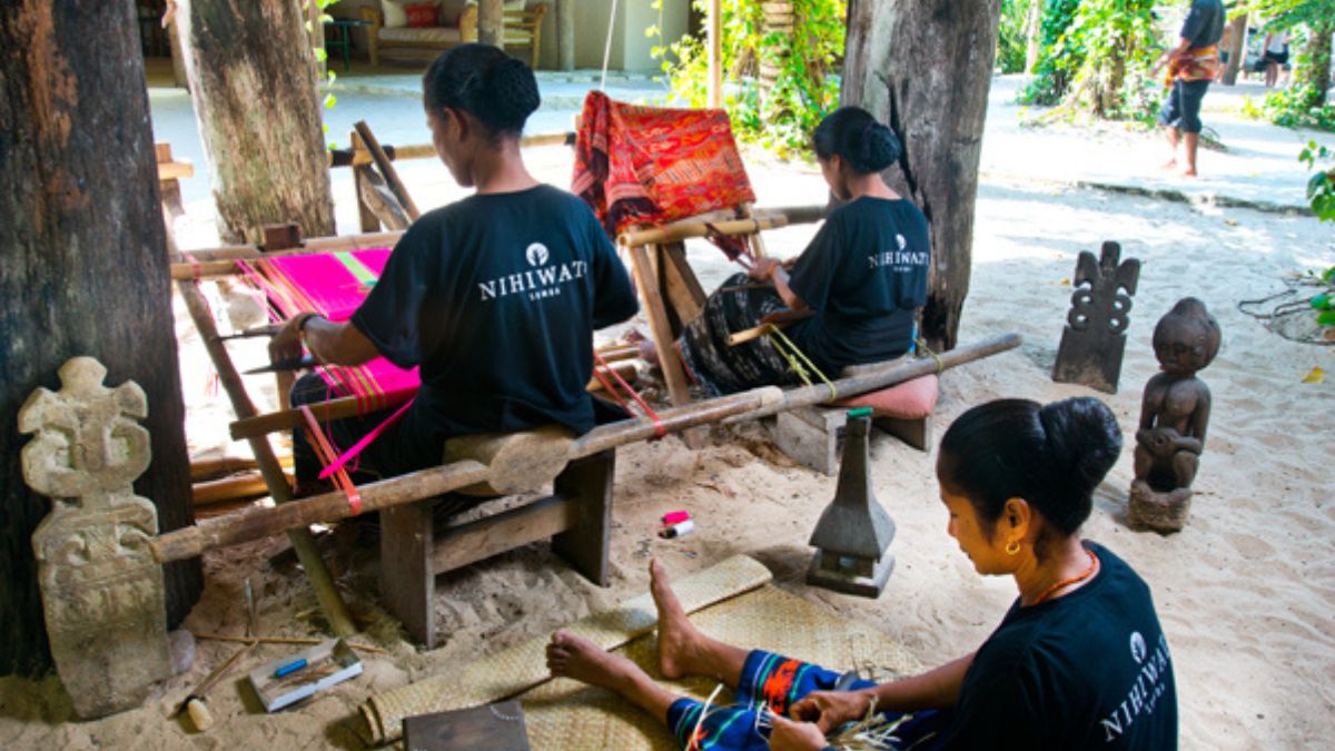 Two women on benches and one on the ground weave traditional designs in Indonesia